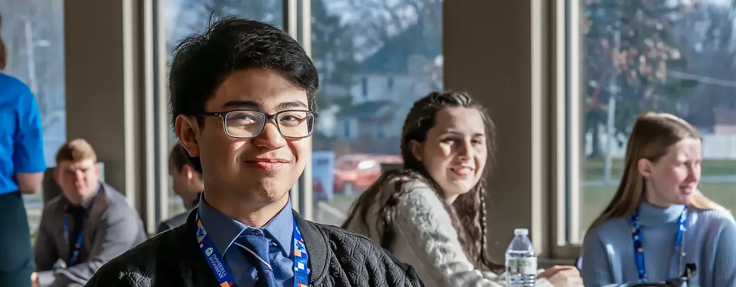 A young man sitting at a table with other students in the background in a classroom setting.